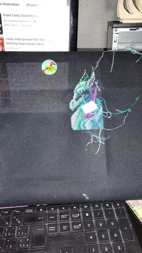 A close-up photo shows a partially completed cross-stitch project on dark fabric, depicting a stylized figure with loose…
