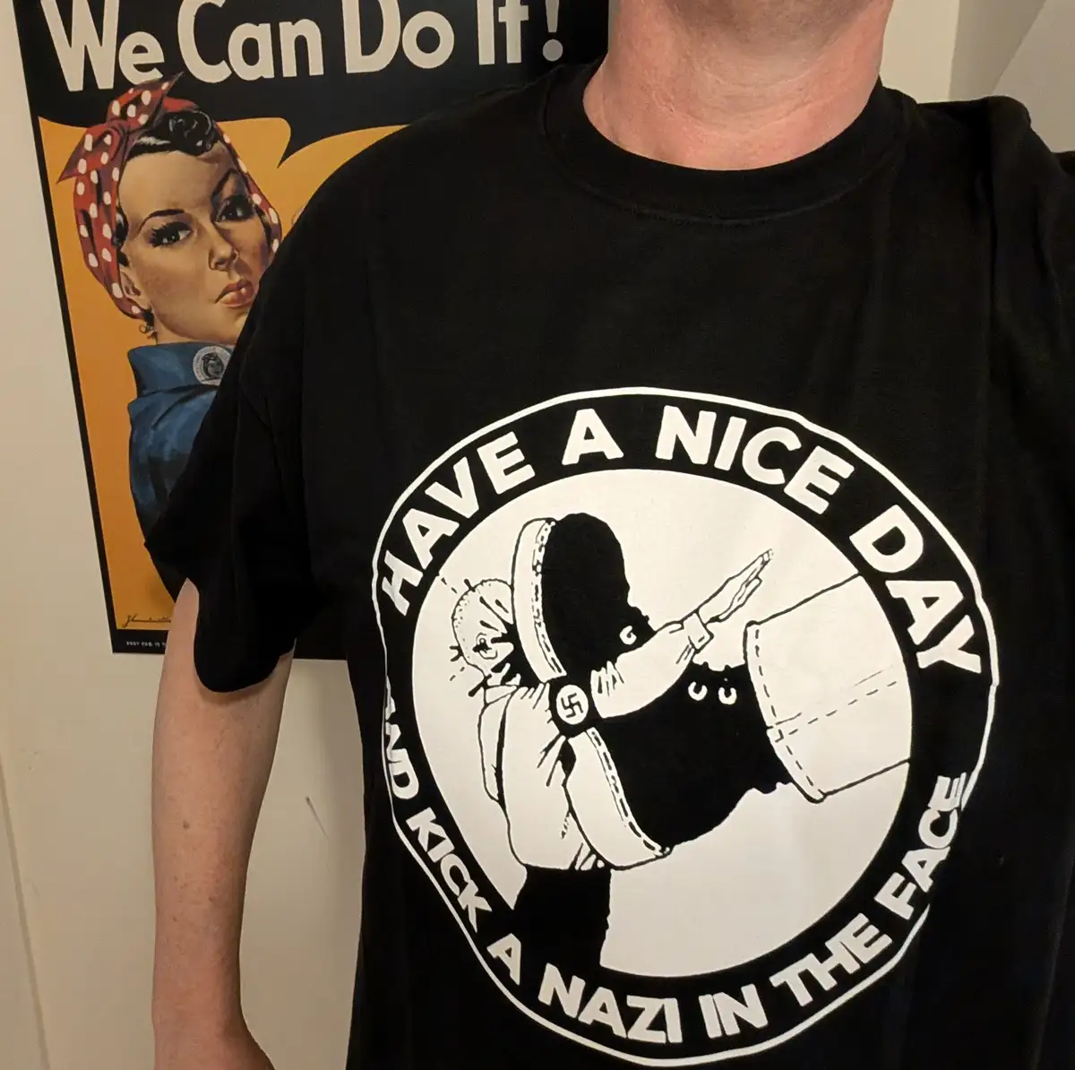 A person wears a black t-shirt with a white circular graphic that reads 'HAVE A NICE DAY AND KICK A NAZI IN THE FACE' around…