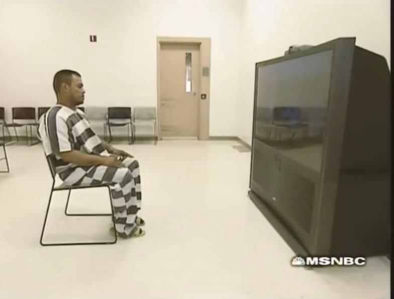 a man in a black-and-white striped prison jumpsuit sits alone in a stark, empty room with rows of chairs and a door in the…