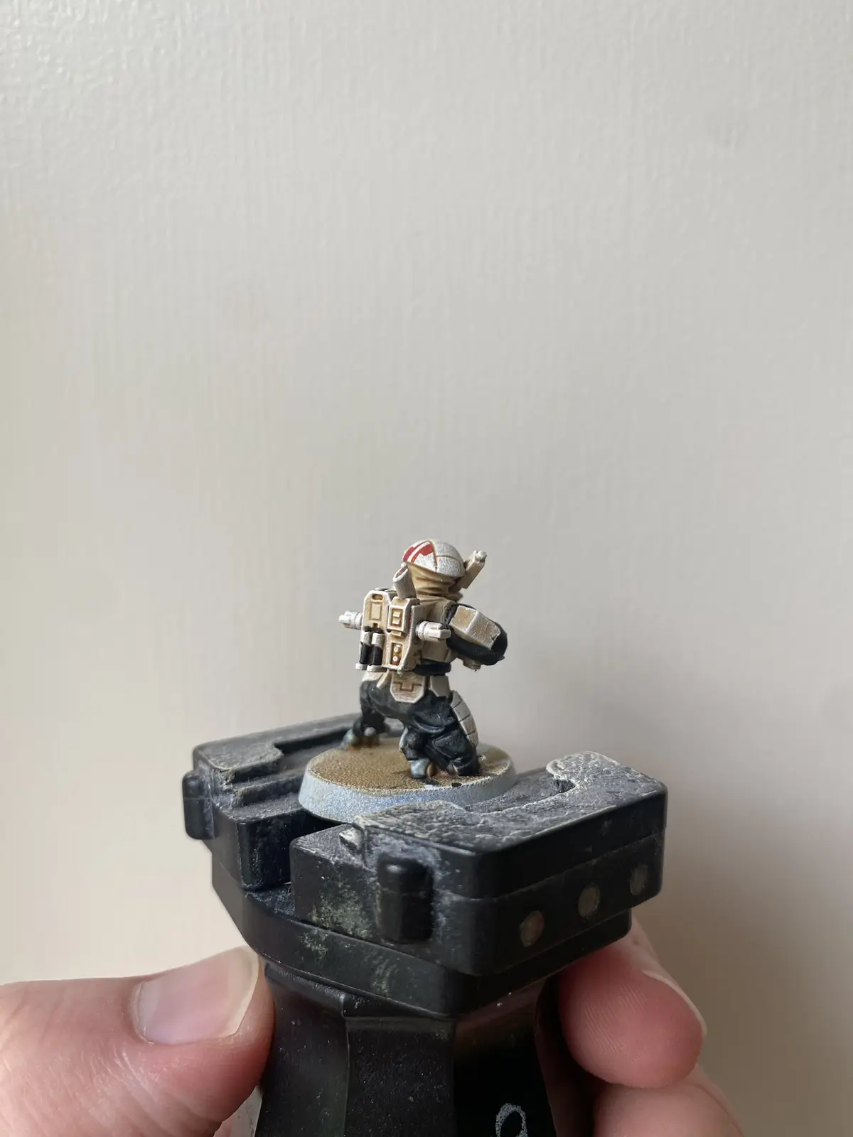 A hand holds a black, worn plastic miniature stand with a painted sci-fi soldier figurine on top. The figurine, wearing a…