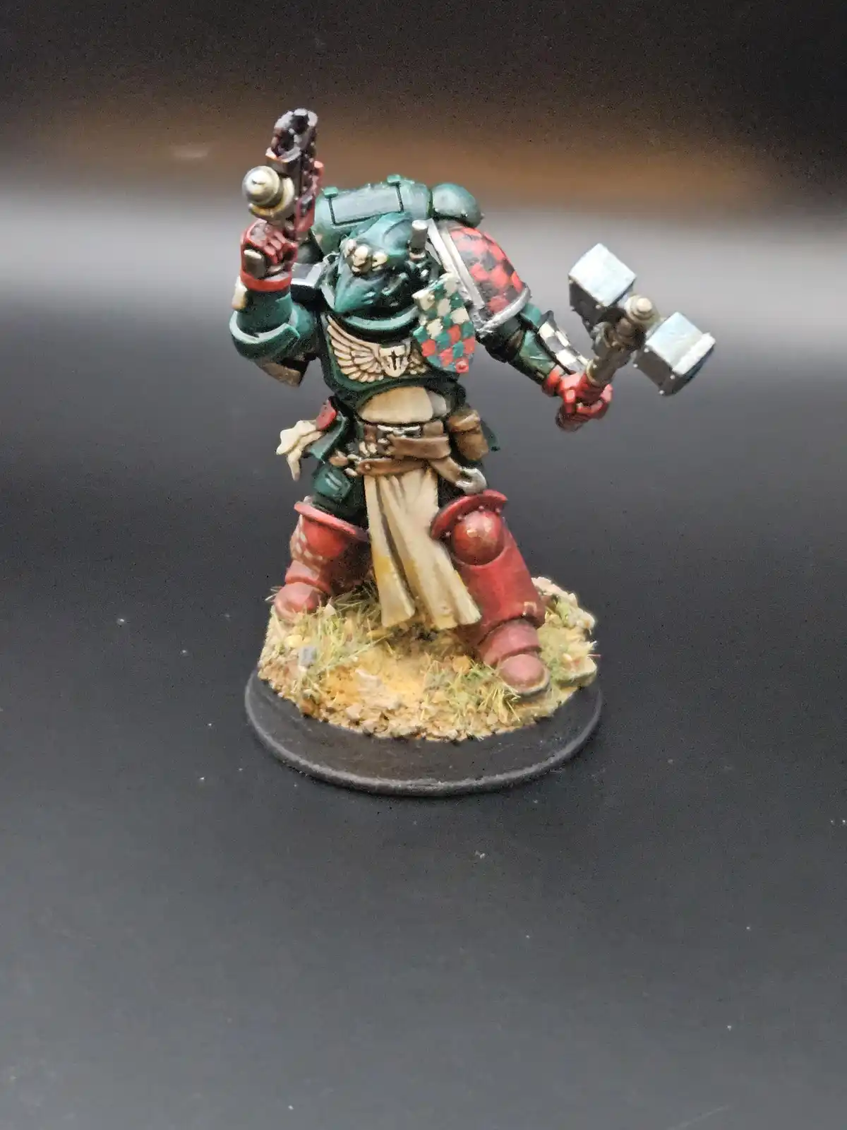 A painted miniature of a green-armored space marine holding a pistol and a hammer stands on a textured base. The overlaid…
