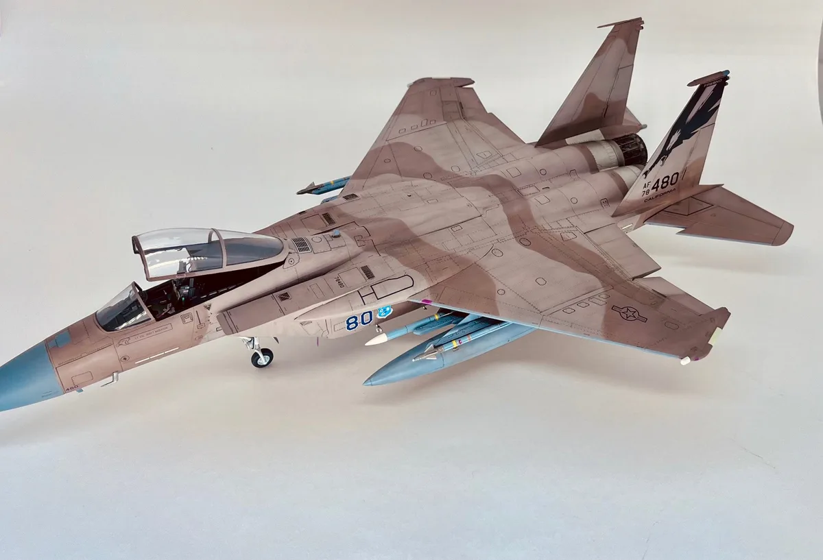 A detailed scale model of a fighter jet, likely an F-15, is shown in a desert camouflage paint scheme with visible markings…