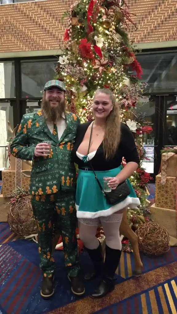 A man with a beard wearing a green suit covered in gingerbread men and 'bite me' text stands beside a woman in a teal skirt…