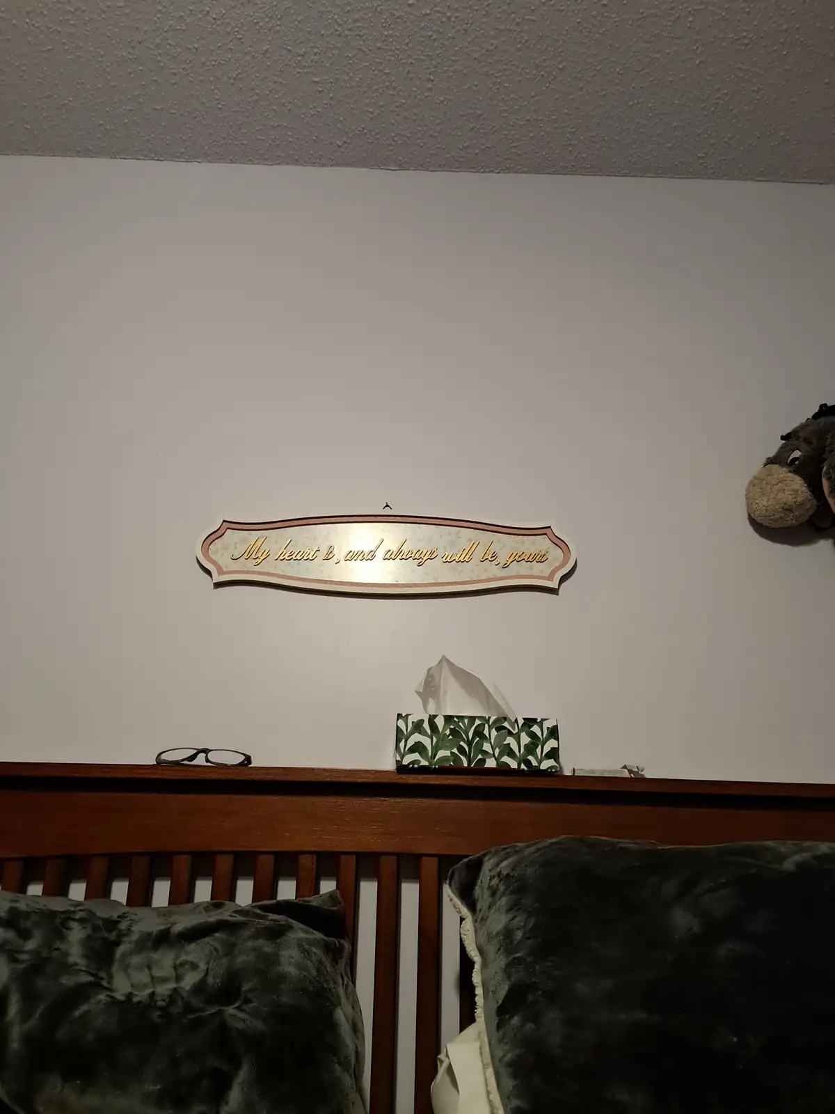 A photo of a bedroom headboard with a decorative sign above it that reads 'My heart is, and always will be, yours'. On the…