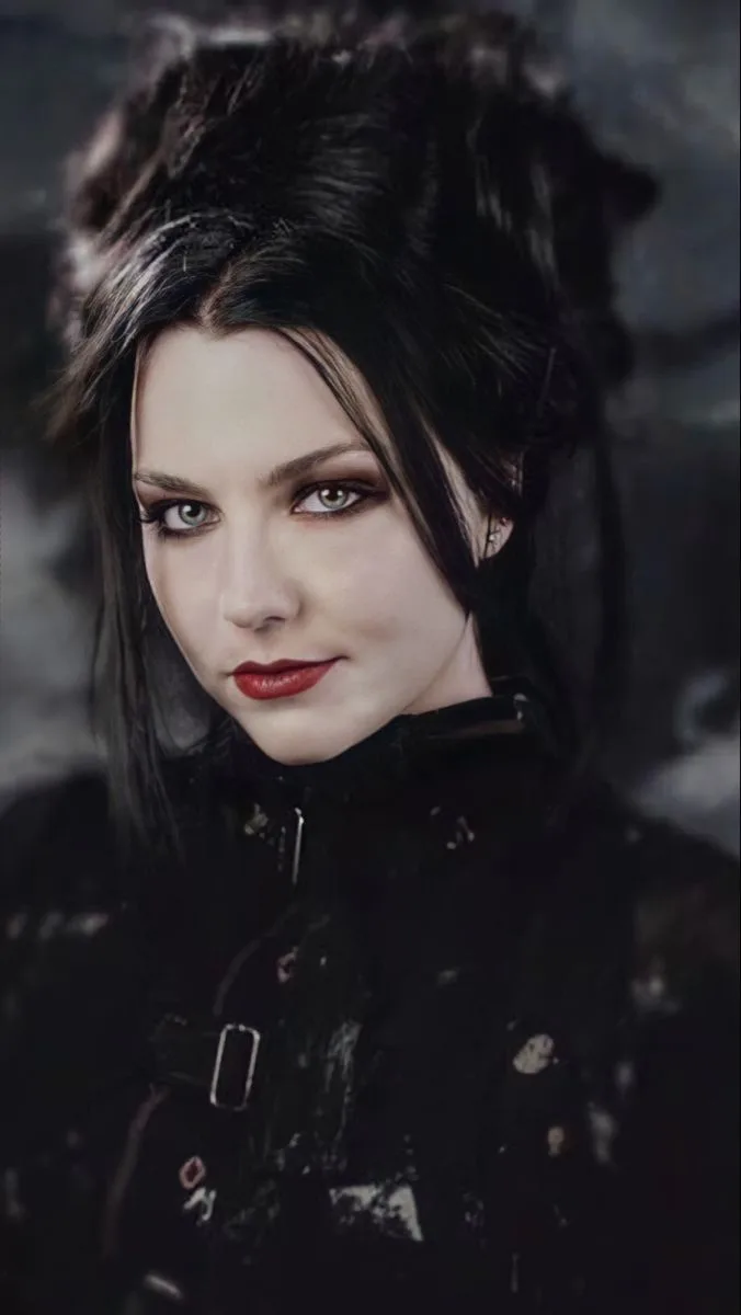 A woman with dark hair, heavy eye makeup, and red lipstick stares directly at the camera with a slight smirk, wearing a…