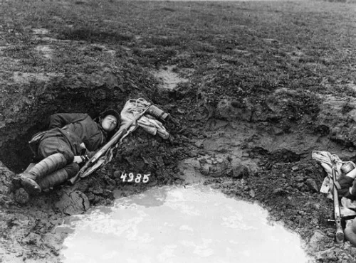 A black-and-white historical photograph shows a soldier lying in a muddy trench, appearing to be asleep or resting. Beside…