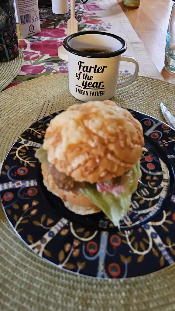 A close-up photo shows a breakfast table with a burger on a patterned plate and a white enamel mug with black rim. The mug…