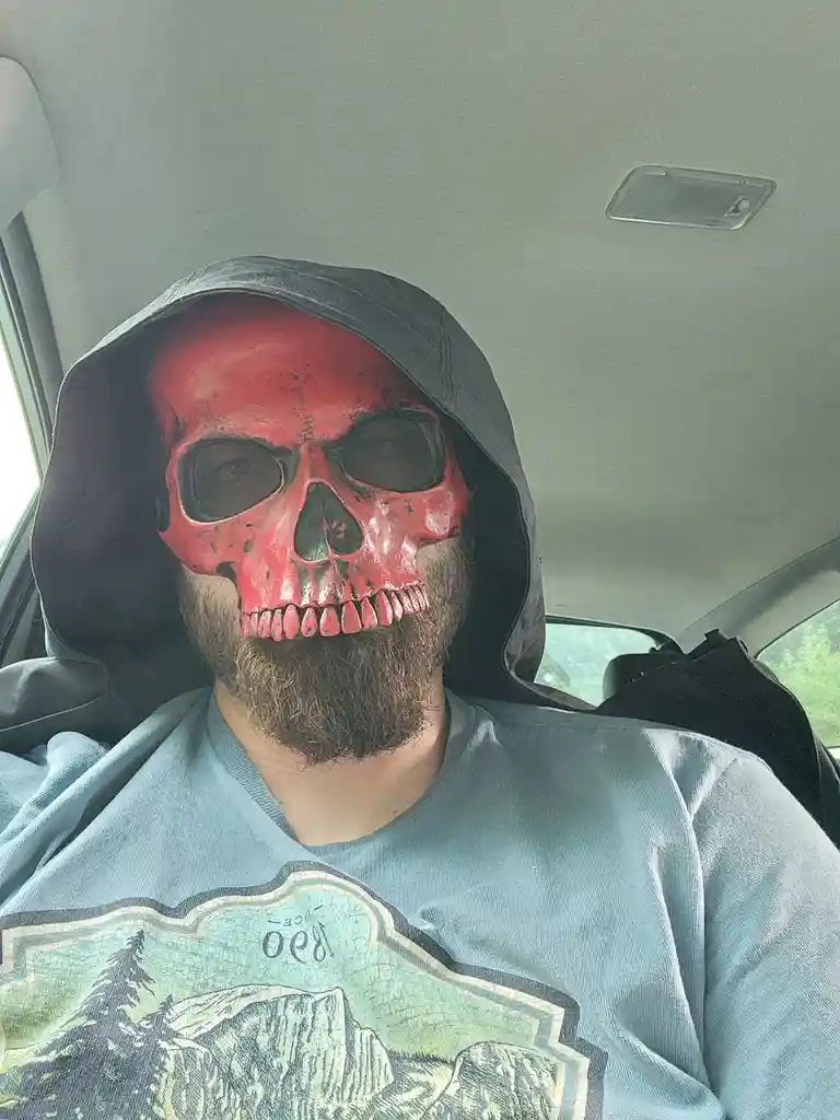 A man with a full beard sits in a car wearing a red plastic skull mask over his face and a black hood pulled up. He wears a…