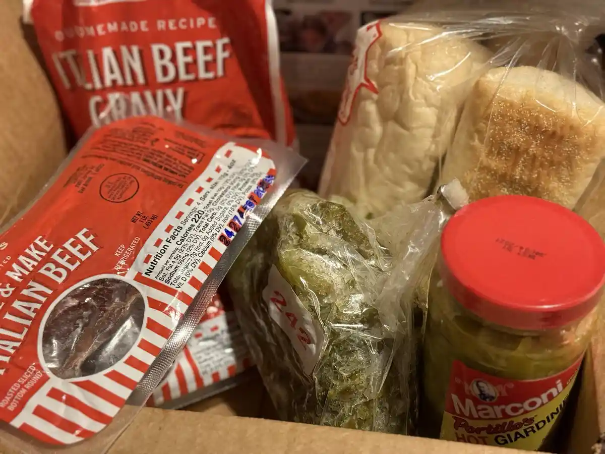 A close-up photo of groceries in a cardboard box, including a package of Italian beef, a jar of Marconi's hot giardiniera, a…