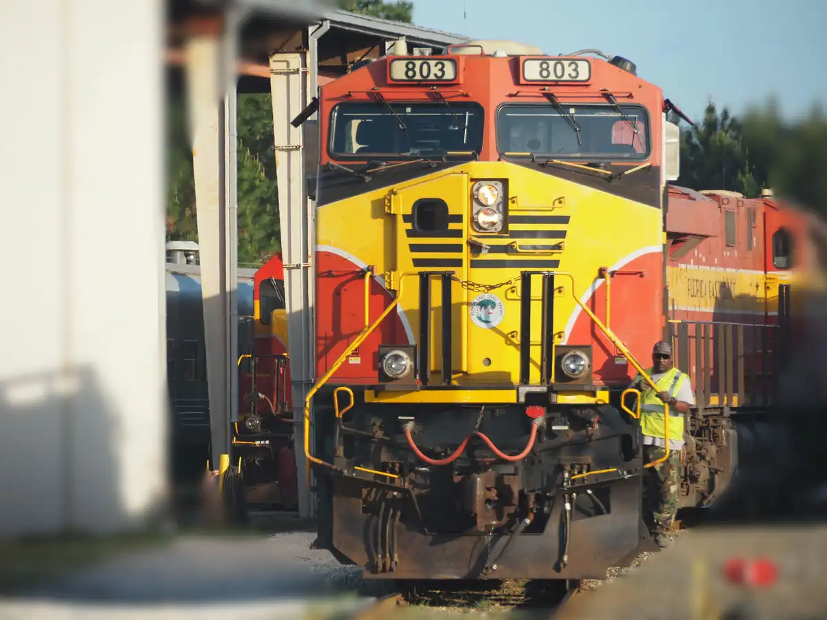 A red and yellow Florida East Coast Railway locomotive, numbered 803, is shown at a rail yard with a conductor in a yellow…
