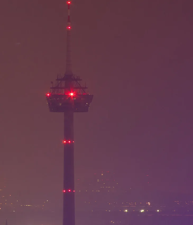 A tall, slender tower with red warning lights at various levels stands against a hazy, purple-tinged night sky. The…