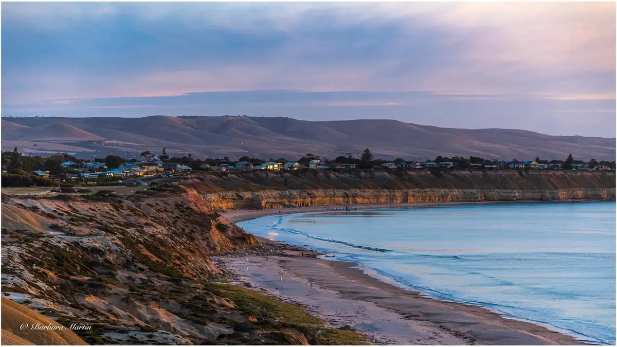 A serene coastal landscape at dusk, featuring a sandy beach, calm blue water, and a row of houses perched on cliffs…