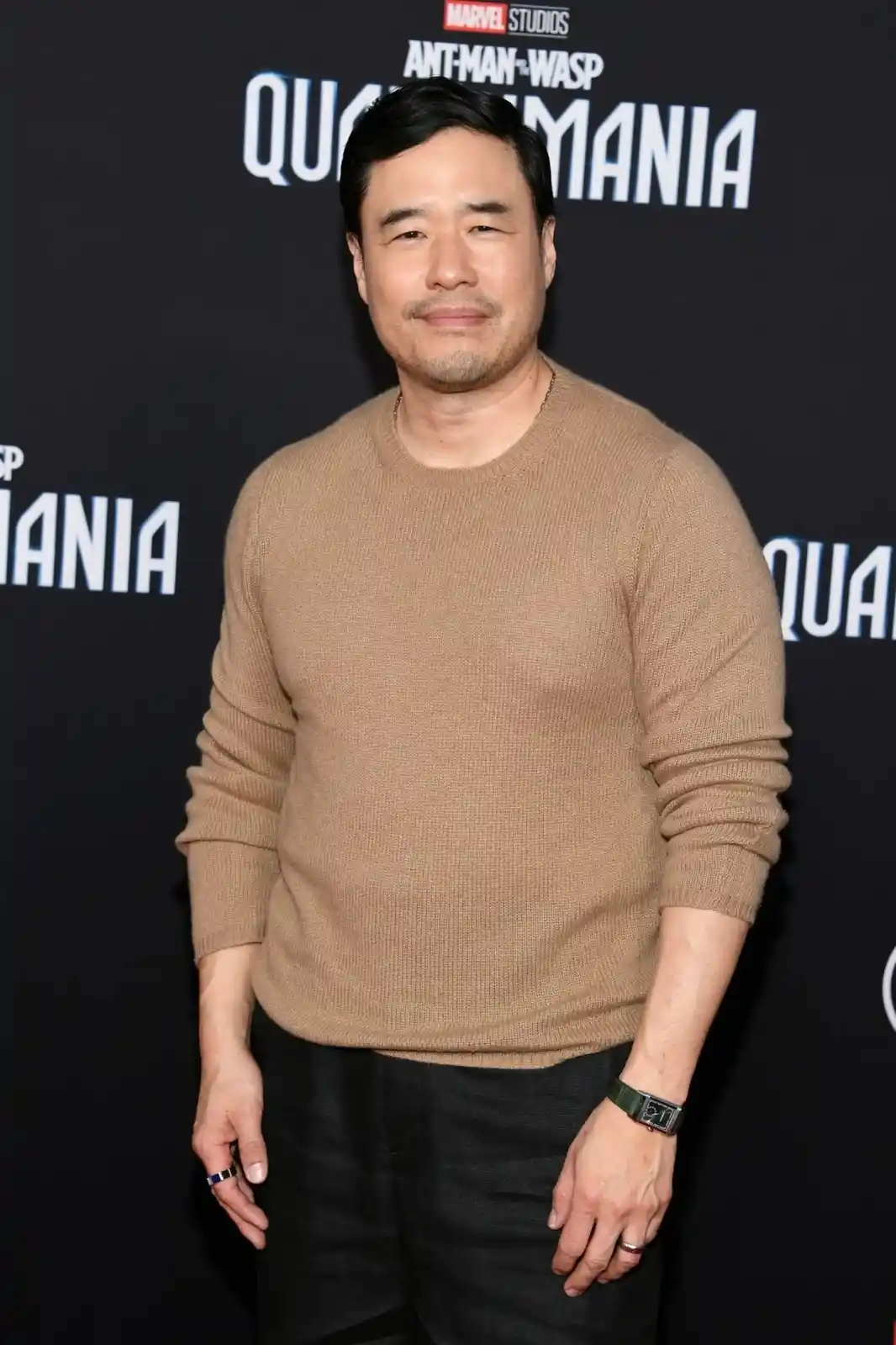 A man in a tan sweater stands at a Marvel Studios 'Ant-Man and the Wasp: Quantumania' premiere, looking unimpressed. The…