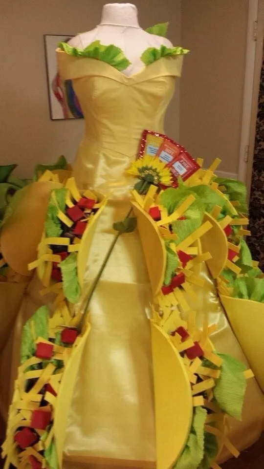 A mannequin wears a yellow satin dress decorated with paper cutouts resembling tacos, complete with green paper lettuce, red…