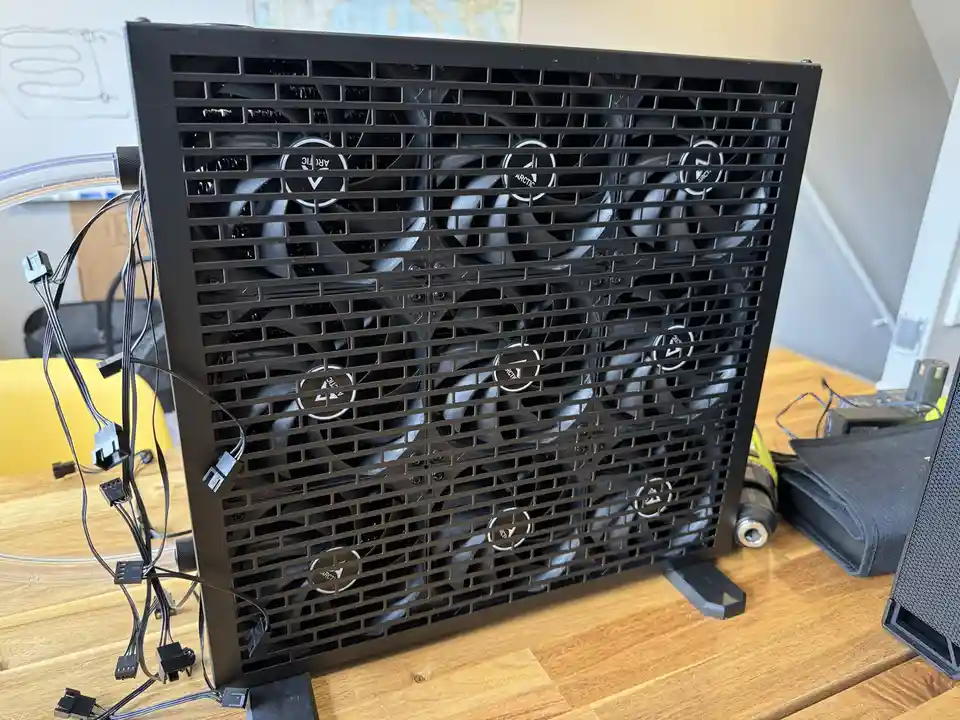 A large black computer case with a grid of 12 visible fans behind a protective grille, sitting on a wooden desk with cables…
