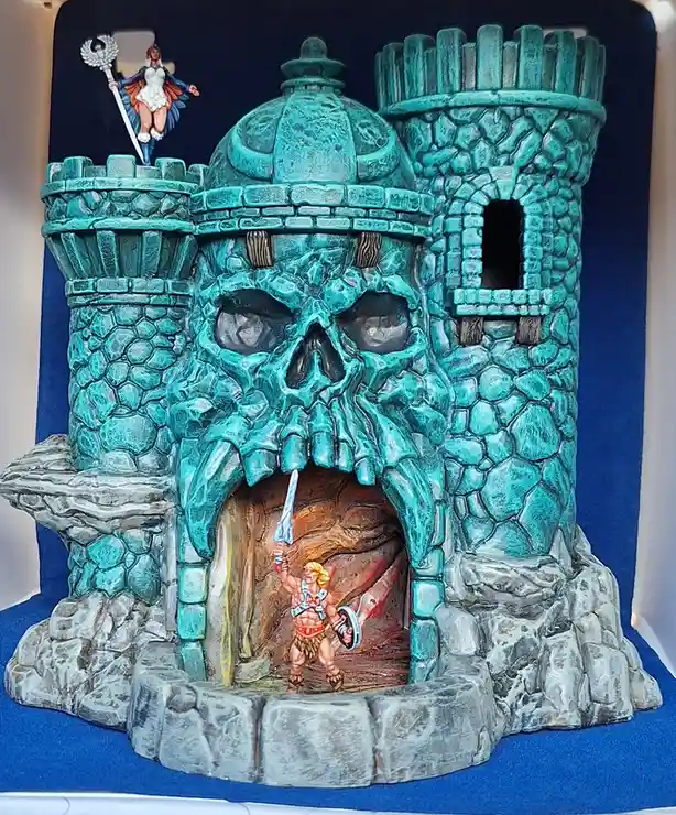 A plastic toy diorama depicts a teal-colored castle shaped like a giant skull with a cave entrance. Inside the cave, a…