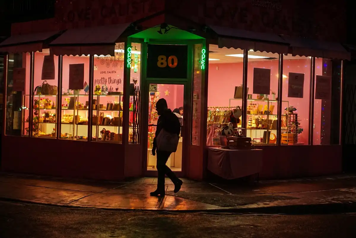 A nighttime photo of a brightly lit pink boutique named 'Love, Calista' with neon 'Open' signs and displays of jewelry and…