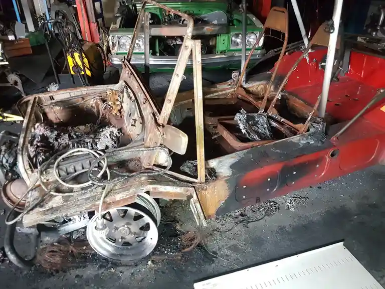 A severely burnt-out red car frame sits in a garage, its interior charred and melted, with only the metal skeleton and one…