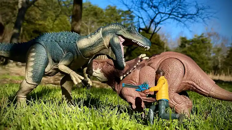 A toy action figure of a man in a yellow jacket holding a blue gun is positioned in grass between two large dinosaur toys…