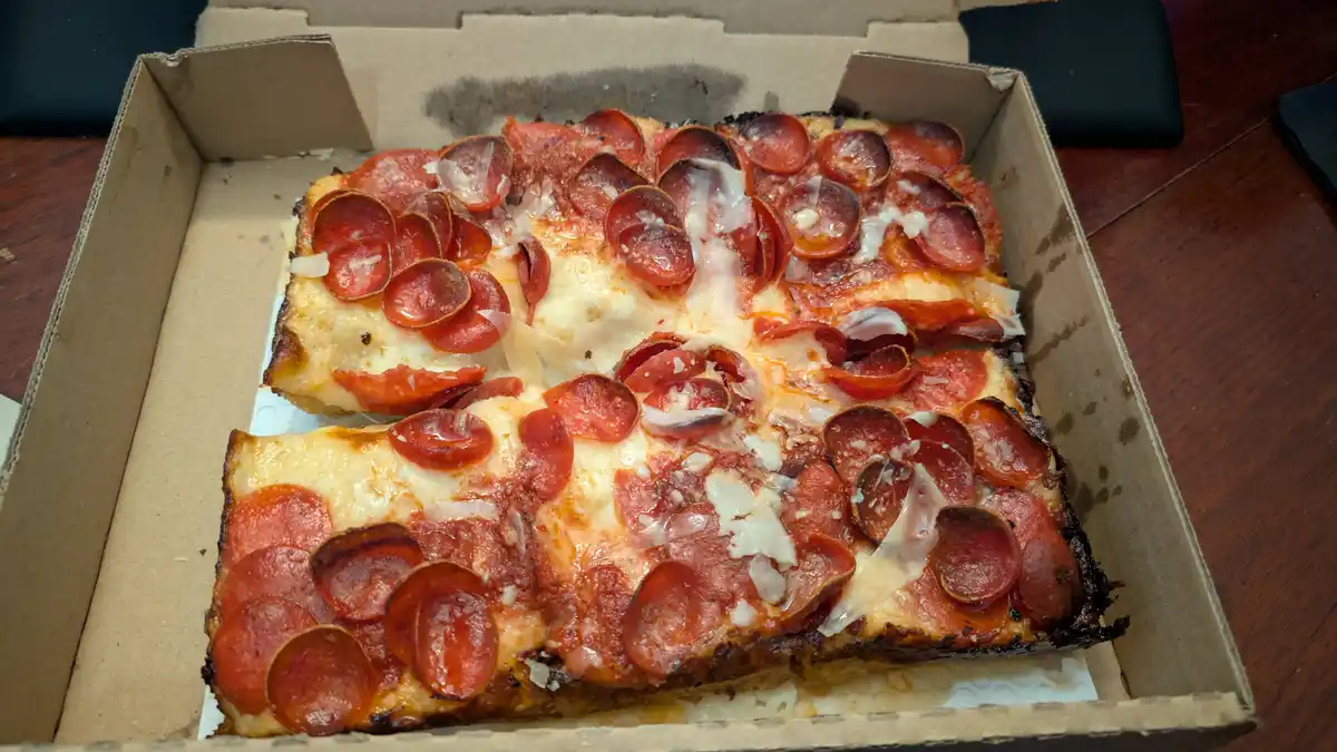 A square pepperoni pizza in an open cardboard box, with one slice missing and the remaining pieces showing melted cheese and…