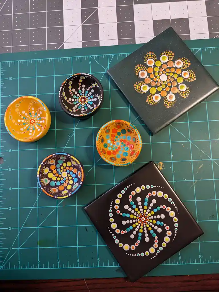 Several small, hand-painted ceramic bowls and square canvases with intricate, colorful dot patterns resembling mandalas are…