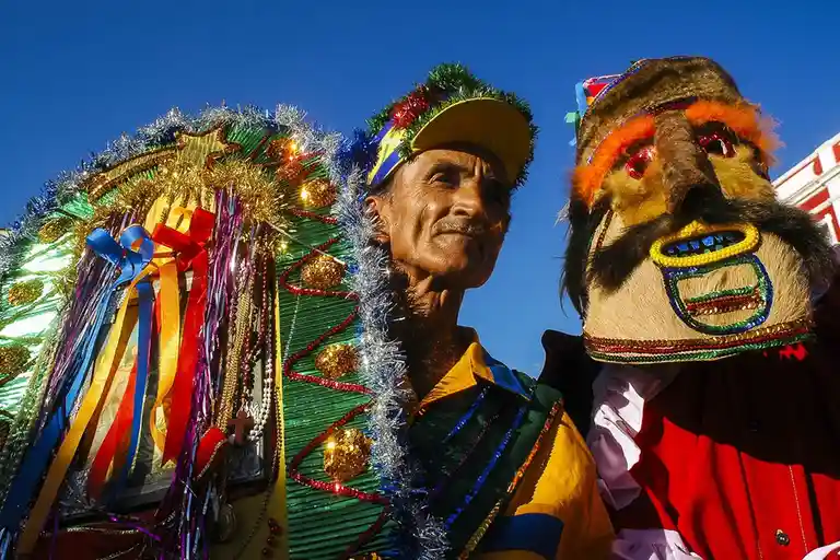 A low-angle photo shows an elderly man in a colorful, tinsel-covered costume with a festive hat, standing beside a person in…