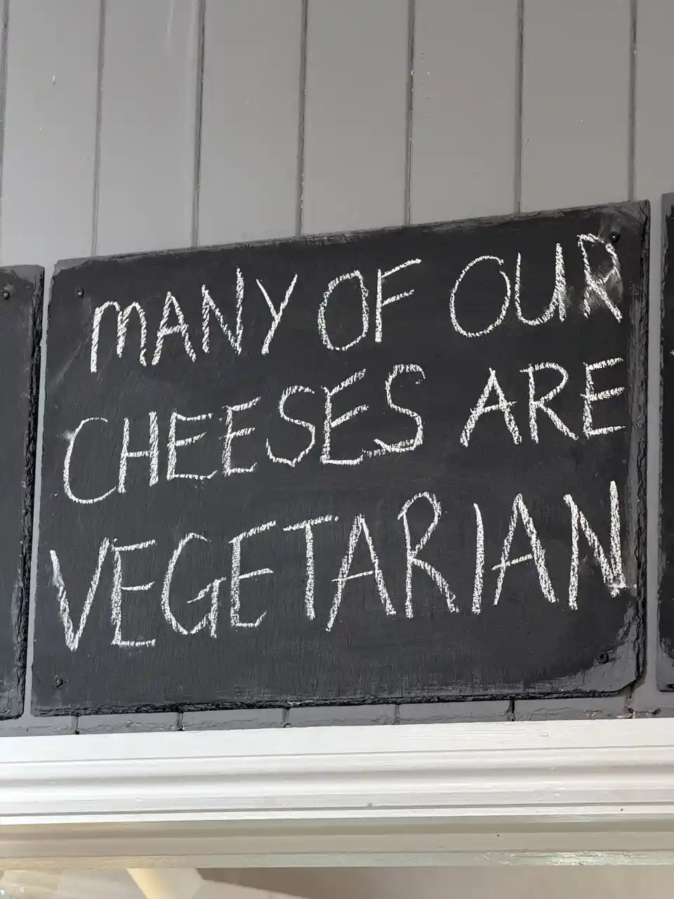 A close-up of a dark slate sign with white chalk handwriting that reads 'many of our cheeses are vegetarian', mounted on a…