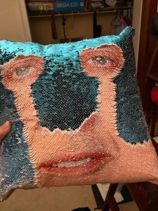 A person holds up a square pillow covered in blue and peach sequins that form a distorted, pixelated face resembling a human…