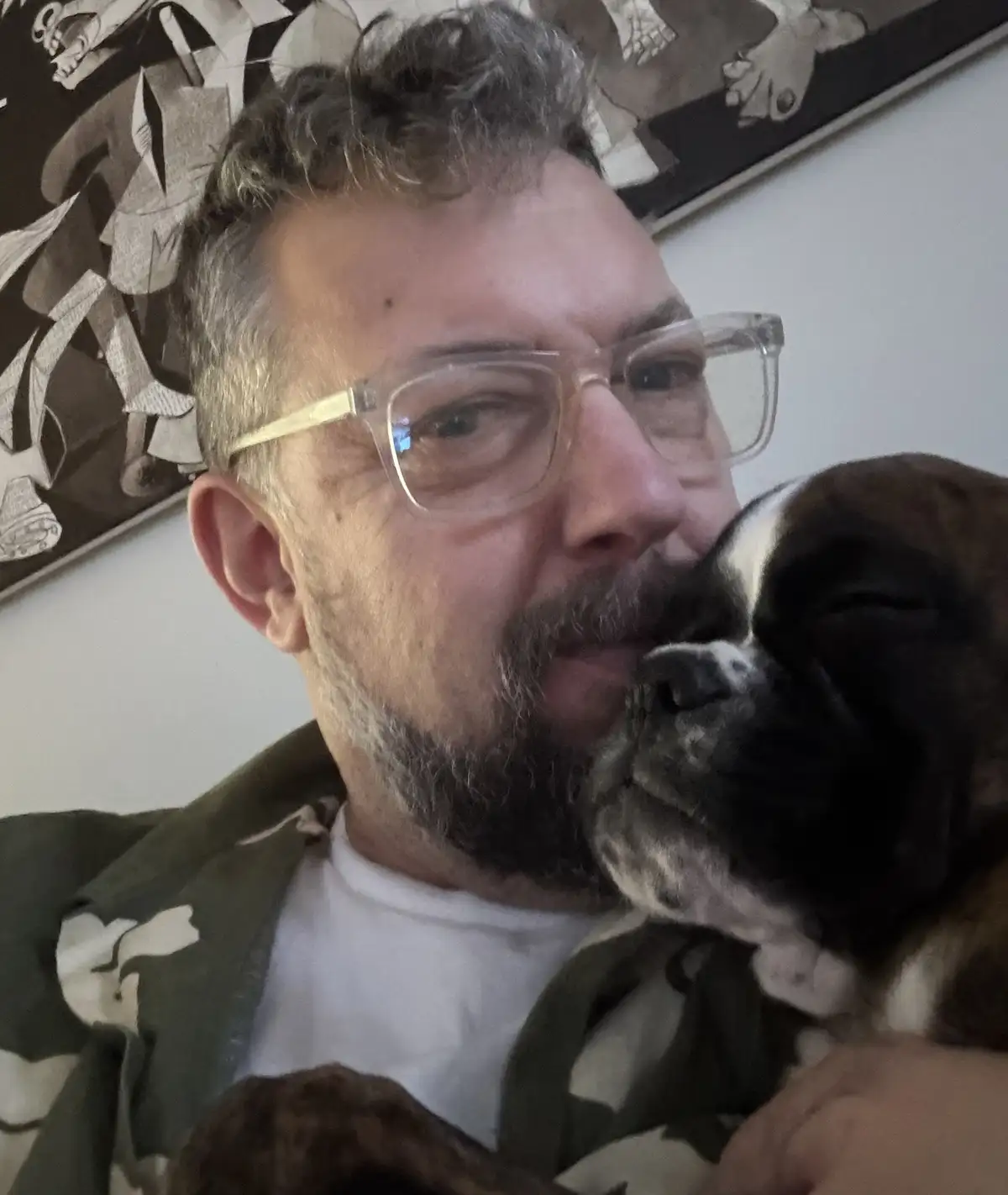 A man with glasses and a beard is holding a black and white dog that is resting its head on his cheek. The man is looking at…