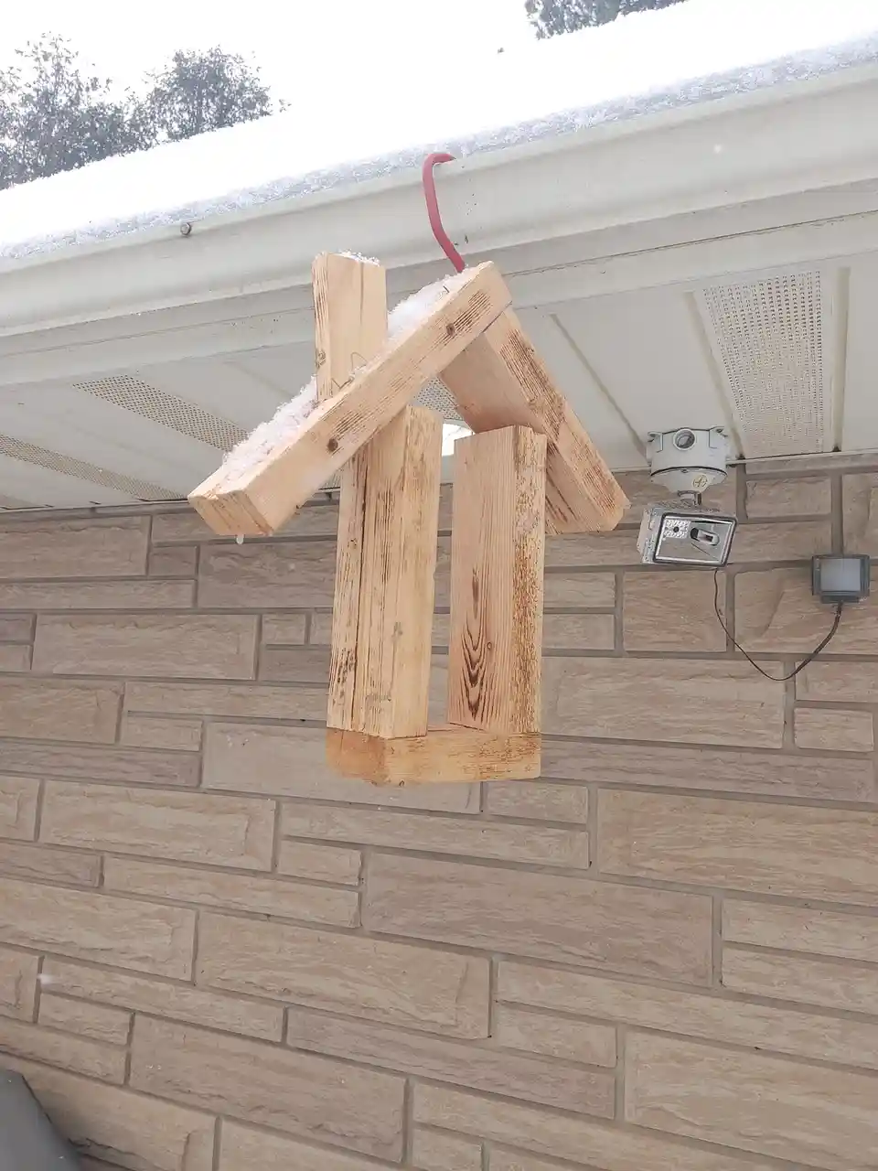 A crudely constructed wooden birdhouse, made from thick planks and hanging by a red hook, is covered in snow and positioned…