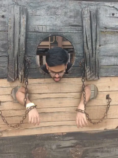 A man with glasses and a beard is shown in three panels, each depicting him in a wooden stocks with chains around his…