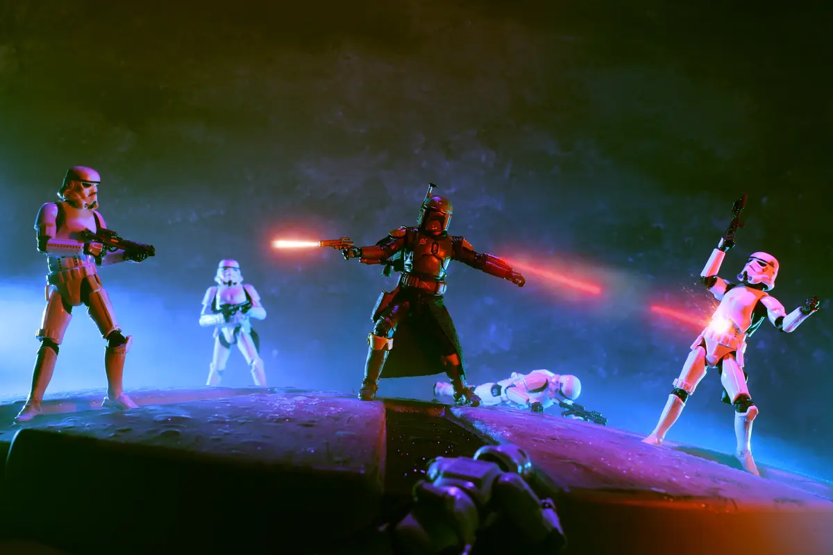A staged photo of Star Wars action figures: a central Boba Fett figure fires red laser beams from dual blasters, flanked by…