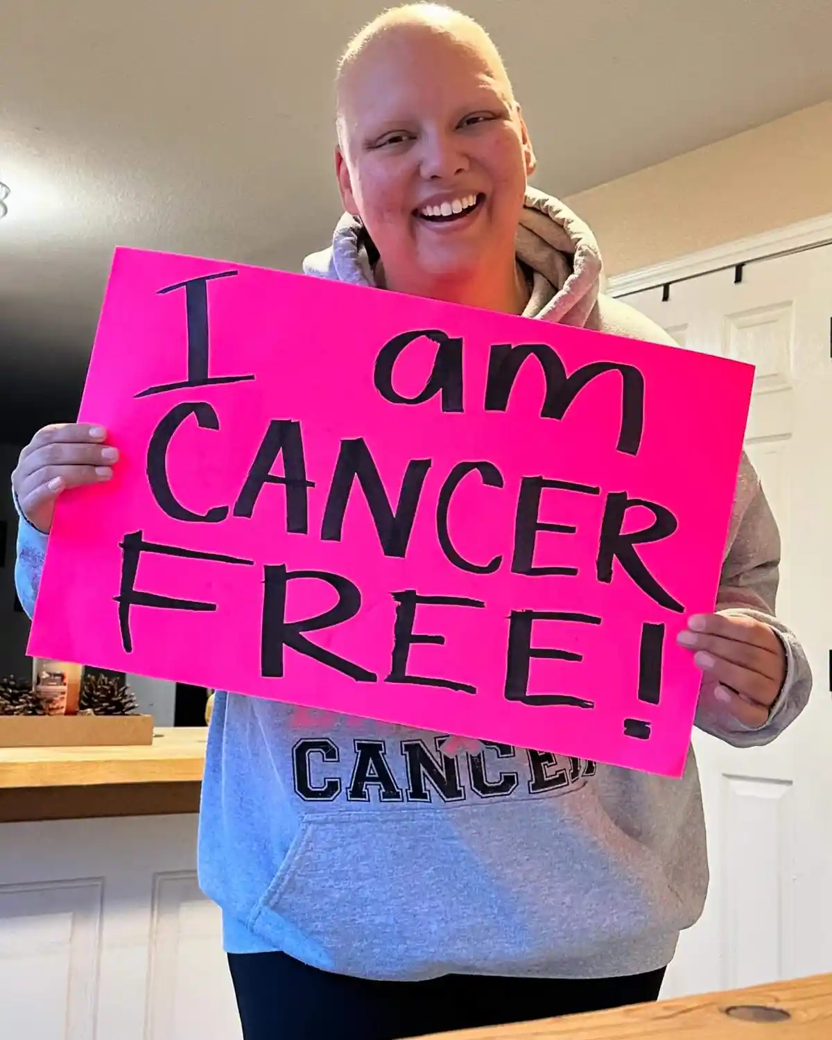 A smiling person with a shaved head holds a bright pink sign that reads 'I am CANCER FREE!' in bold black marker. They are…