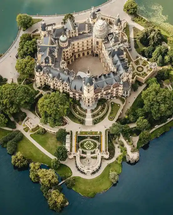 An aerial view of a grand, ornate castle complex situated on a lush, tree-surrounded island in the middle of a large body of…