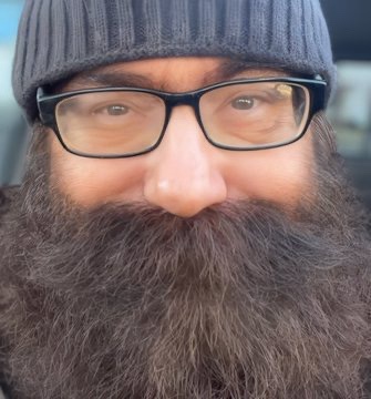 A close-up selfie of a man with a thick, dark brown beard, wearing black-rimmed glasses and a gray knit beanie. The text…