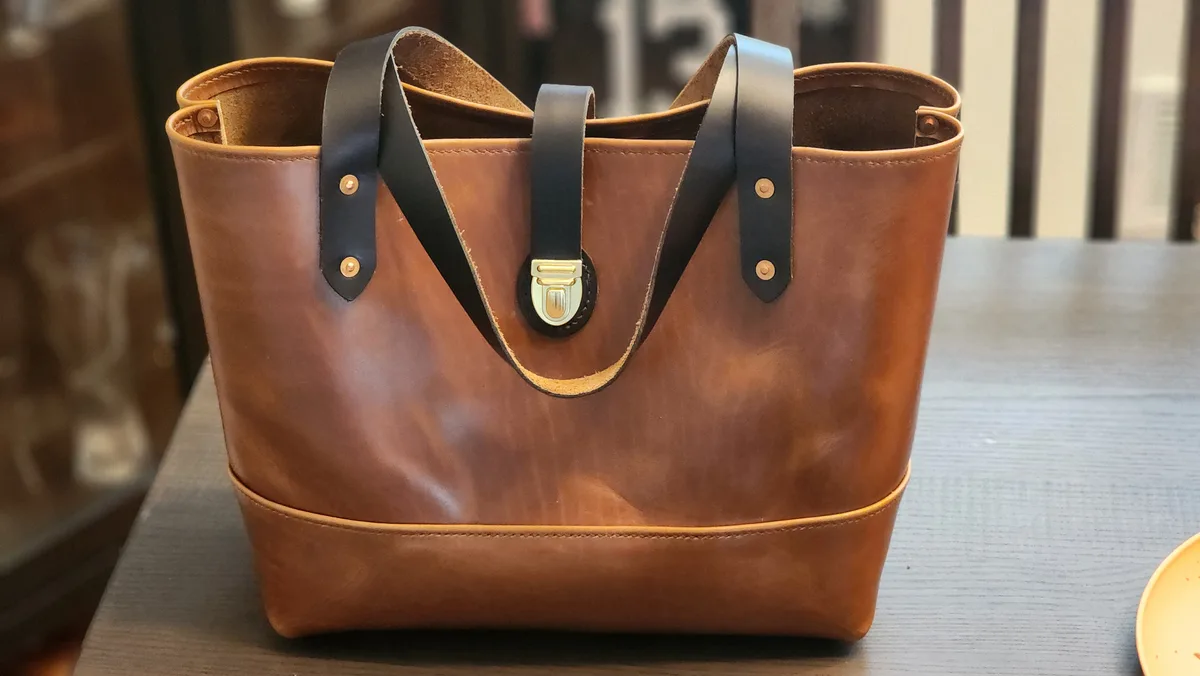 A brown leather tote bag with black straps and gold hardware sits on a wooden table. The overlaid text reads 'i bought this…