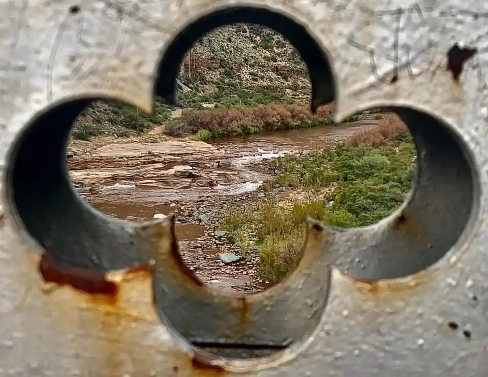 A rusty, weathered metal surface with a quatrefoil-shaped hole frames a view of a muddy river flowing through a rocky…