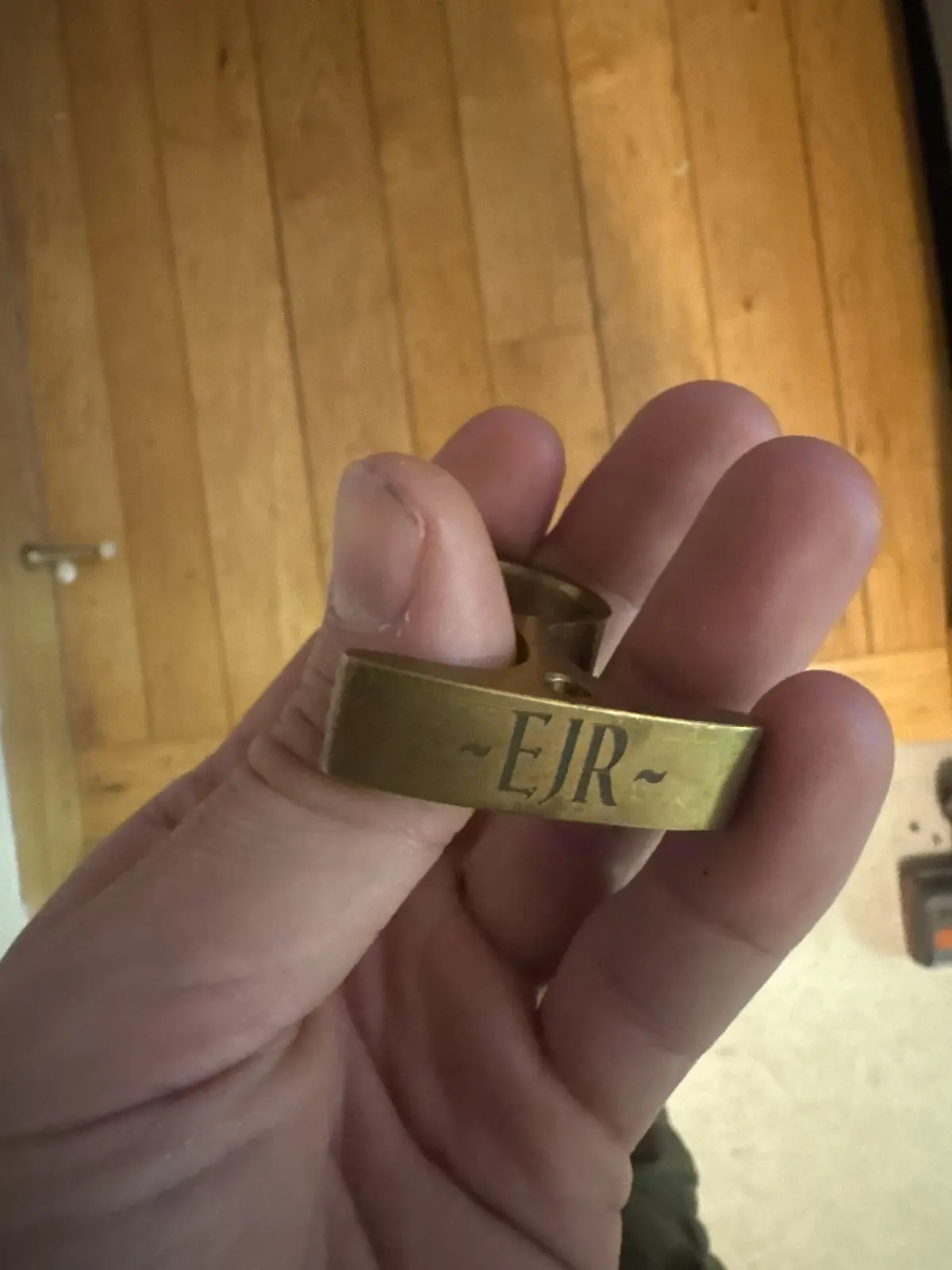 A hand holds a brass finger ring with the engraved initials '~EJR~' against a wooden floor background. The ring is a simple…