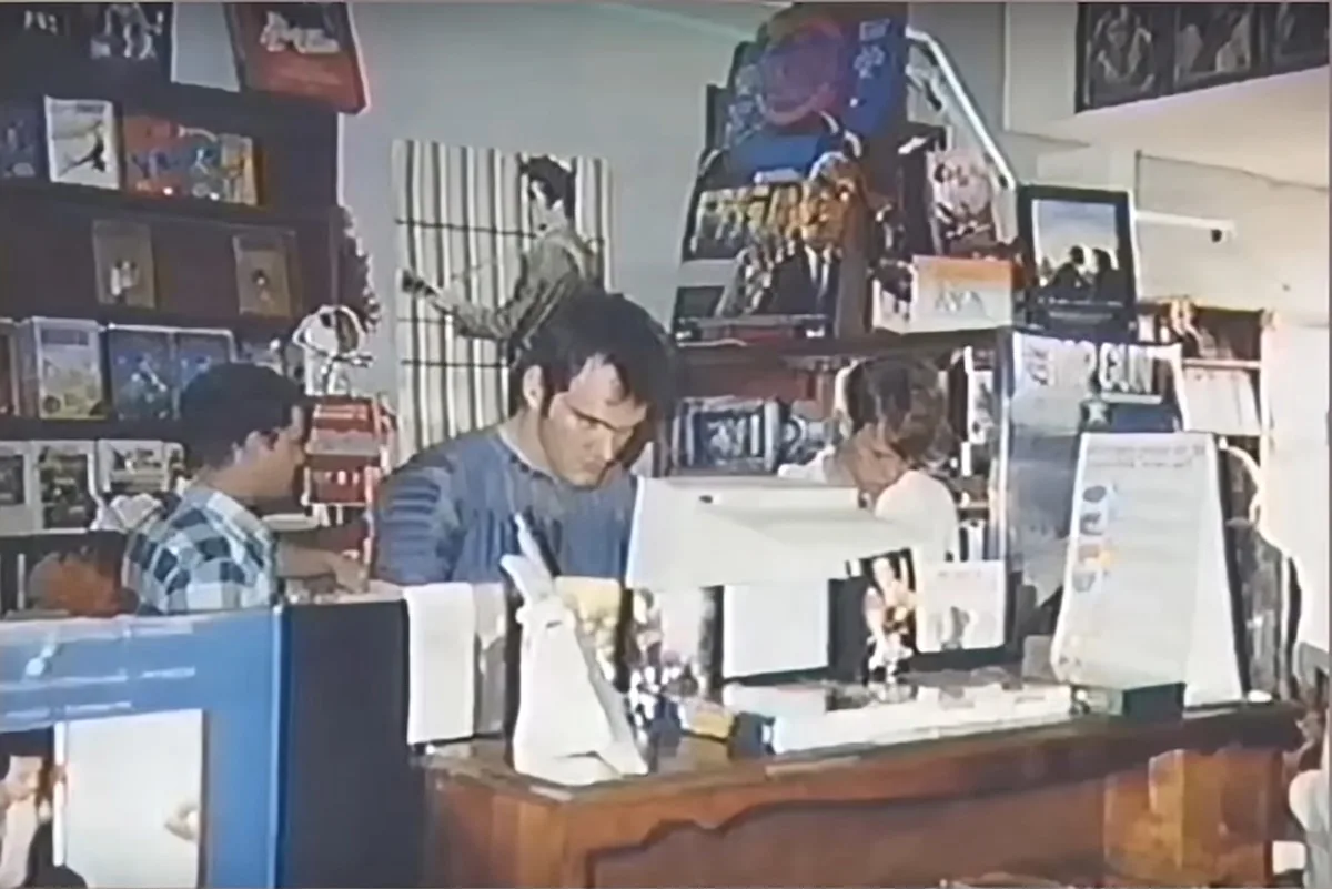 A grainy security camera image shows three men in a store, one in a plaid shirt facing left, one in a striped shirt looking…