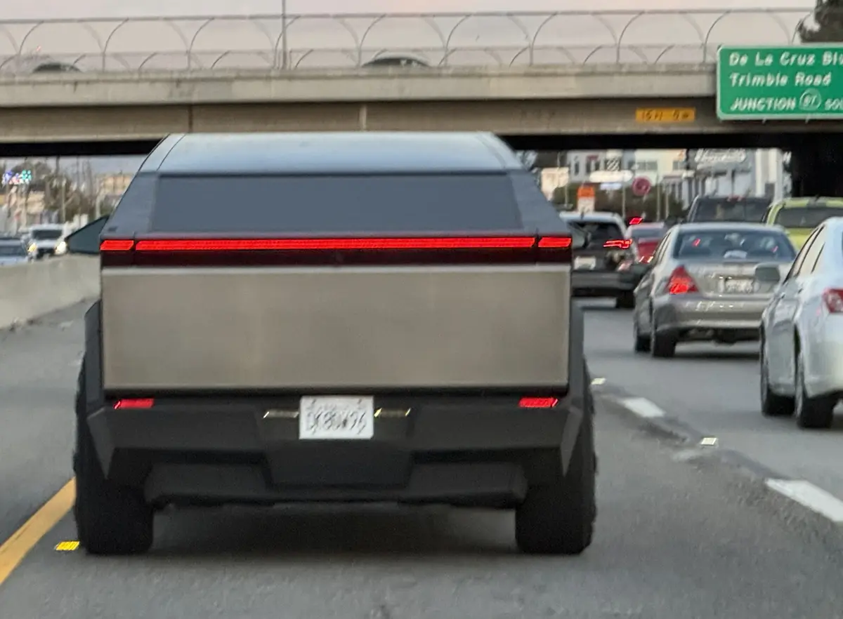 The image shows the rear of a black Tesla Cybertruck on a busy highway under an overpass with a green road sign reading 'De…