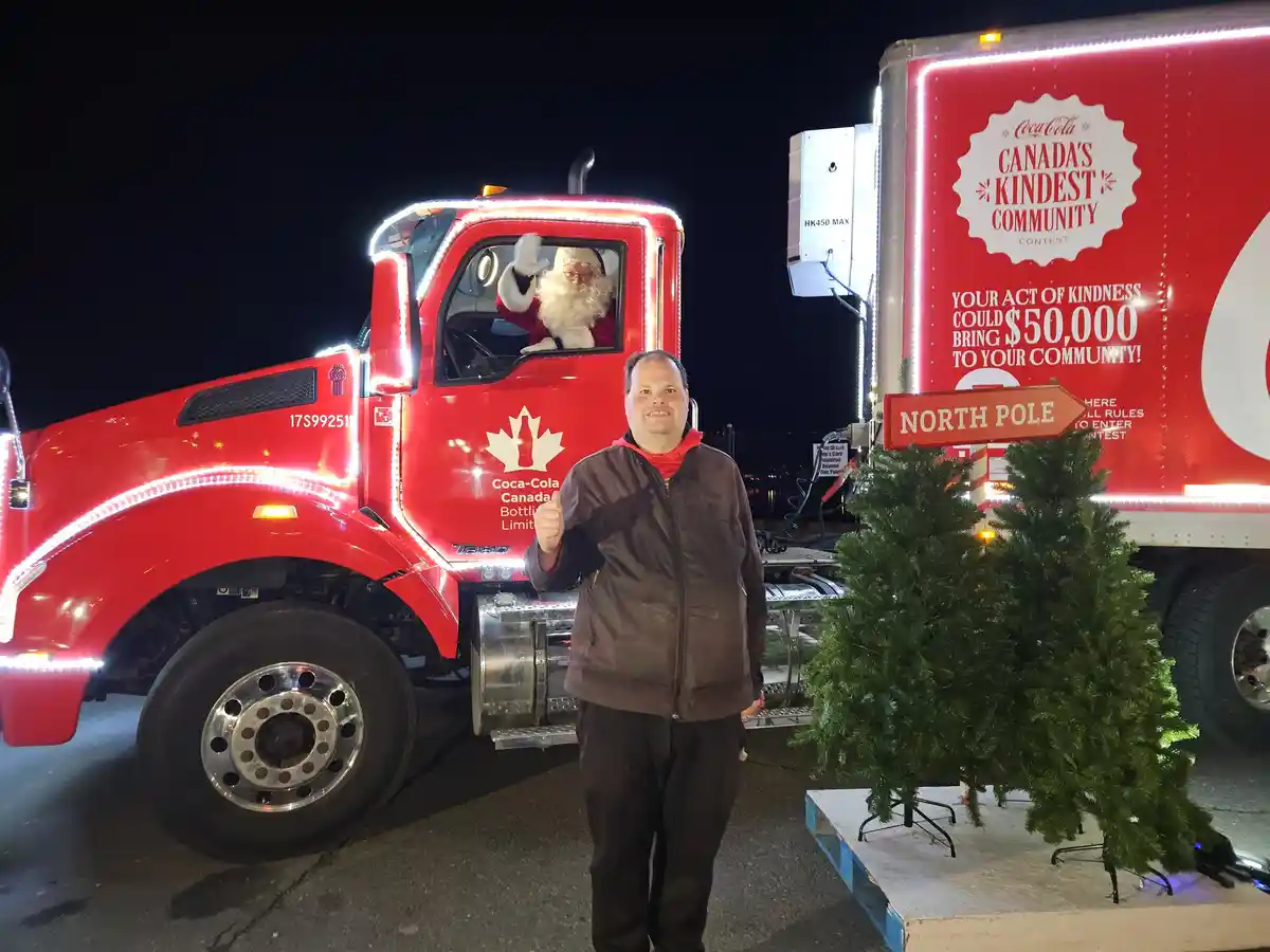 A man stands in front of a red Coca-Cola truck decorated with Christmas lights, giving a thumbs-up. Inside the truck's cab…