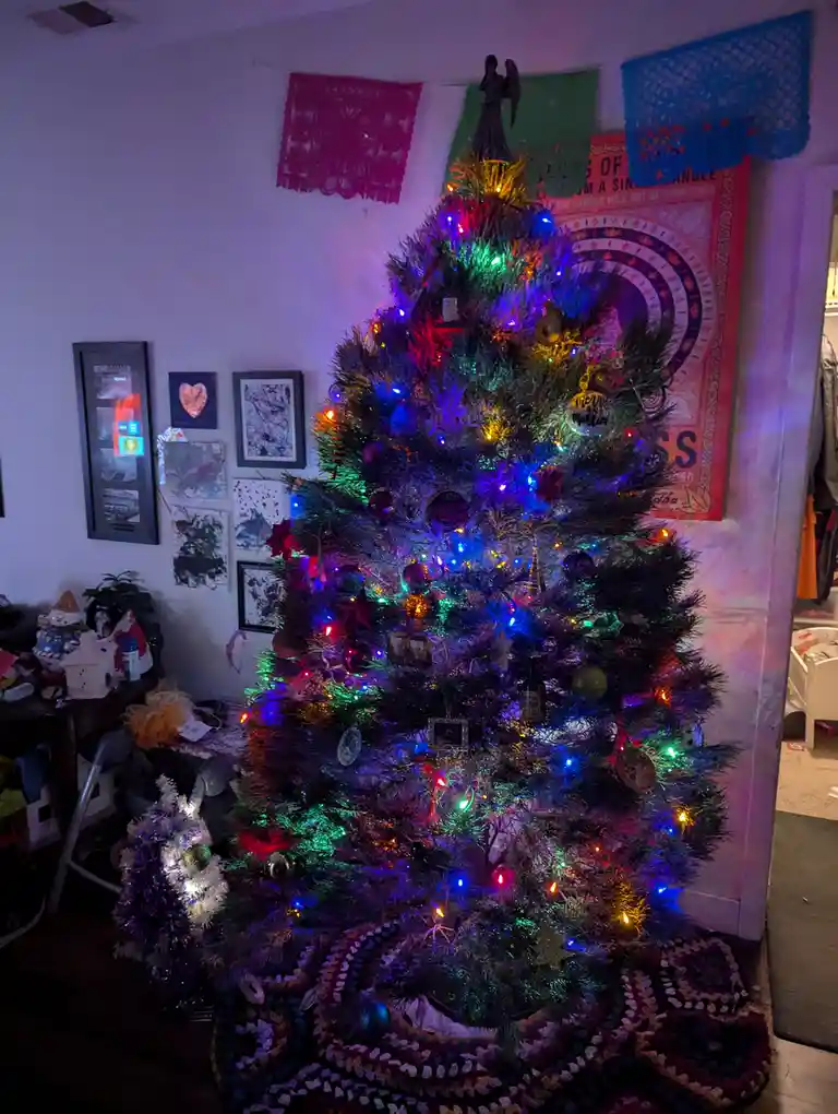 A heavily decorated, multi-colored Christmas tree with blinking lights and various ornaments stands in a cluttered room…