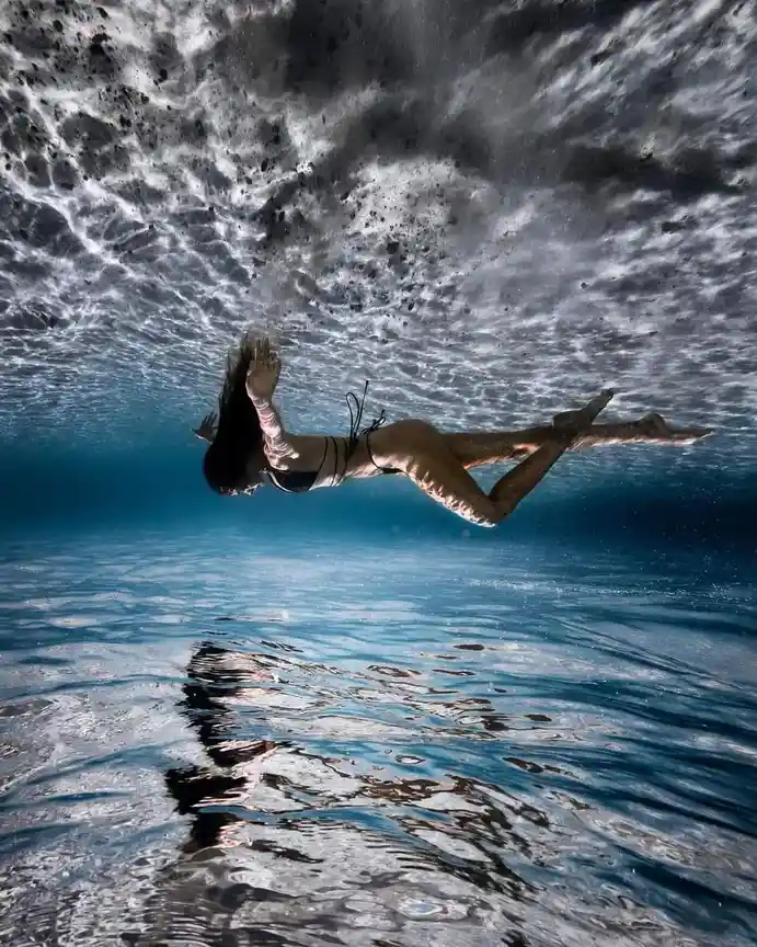 An underwater photograph shows a woman in a black bikini swimming gracefully beneath the surface of clear blue water, with…