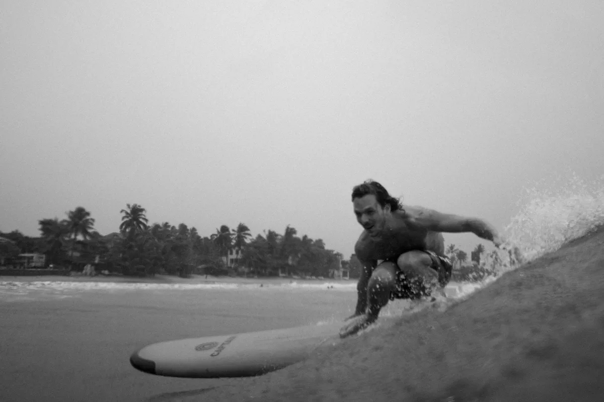 A black-and-white photo shows a shirtless man crouched on a surfboard riding a wave, with palm trees and beach visible in…