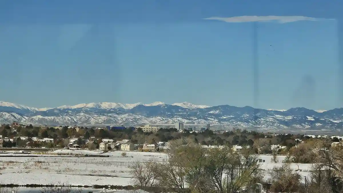 A scenic winter landscape viewed through a window, showing snow-covered fields, residential houses, and distant snow-capped…