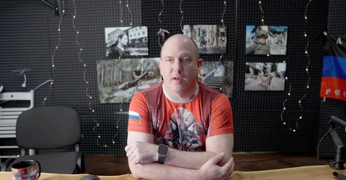a bald man in an orange t-shirt with a Russian flag patch on his sleeve sits at a desk with arms crossed, looking directly…
