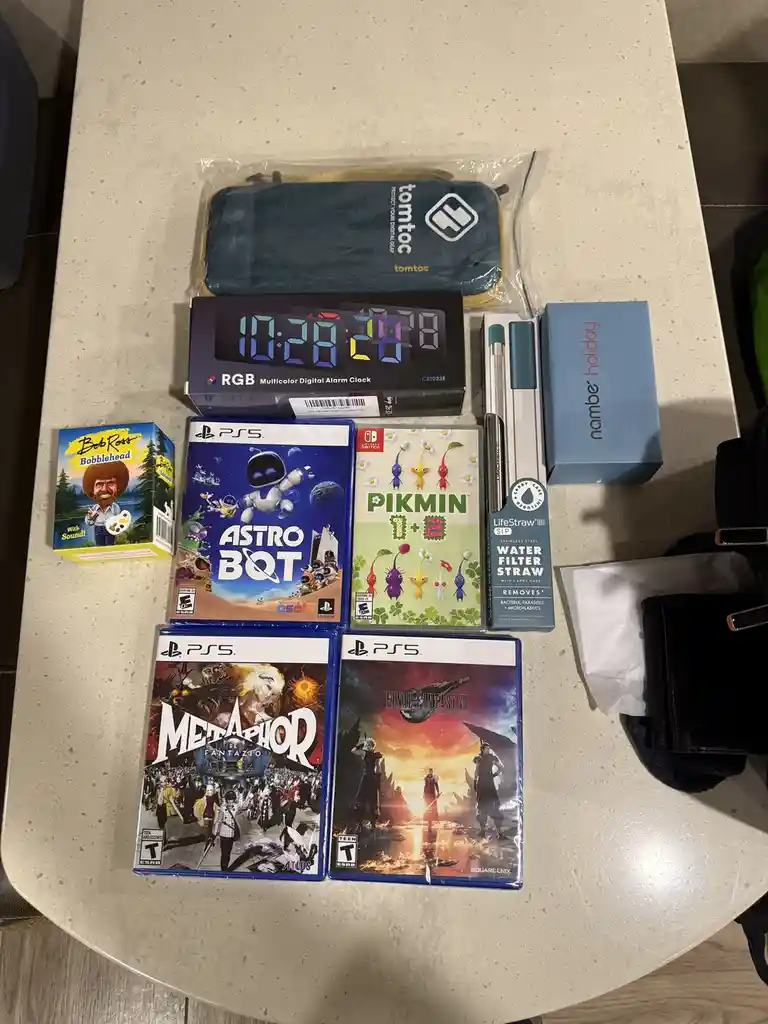 An overhead shot displays a collection of items on a light-colored countertop, including four PS5 game cases (Astro Bot…