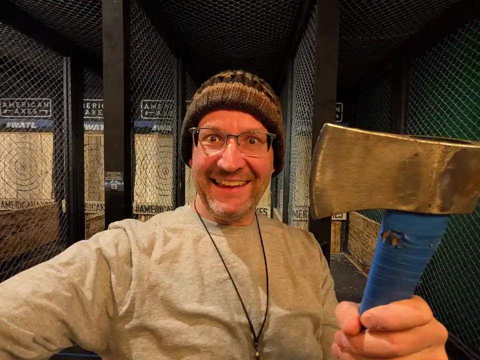 A man with glasses and a knit beanie smiles widely while holding an axe with a blue handle in an axe-throwing facility…