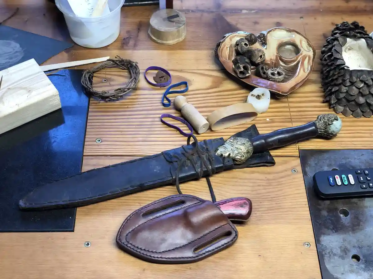A wooden table displays a handmade sword with a decorative brass hilt and a black leather sheath, alongside a brown leather…