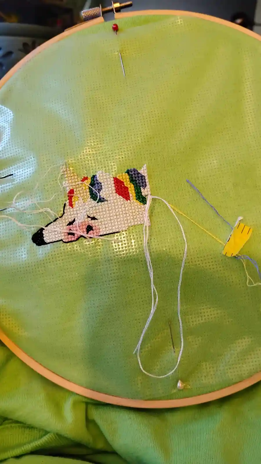A close-up photo shows a partially completed cross-stitch of a unicorn on green fabric held in a wooden embroidery hoop. The…